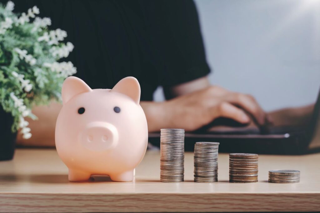 piggy bank and stack of coins with someone using a computer in the background